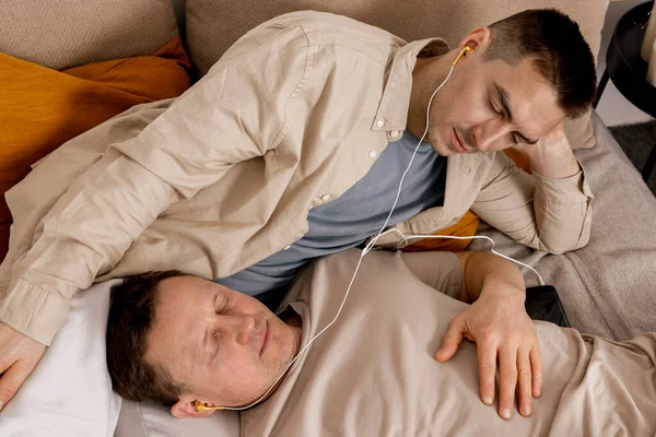 Šťastný gay pár s neformálním oblečením tráví čas doma a poslouchá hudbu se sluchátky. Dva běloši odpočívají. Homosexuální vztahy a alternativní láska. Útulný interiér. — Stock fotografie