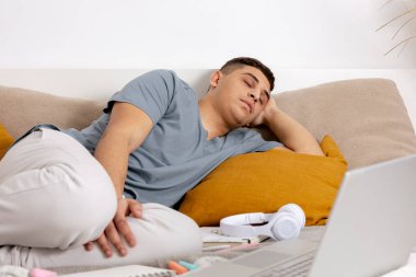 Genç adam ders çalıştıktan sonra çok yorgun ve uykuya daldı. Evde dizüstü bilgisayarla yatağa uzanmış sıradan elbiseli bir adam. E-öğrenme platformunu kullanarak. Uzaktan eğitim. İçerisi rahat. Son teslim tarihi.