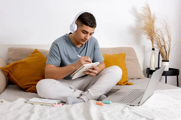 Mladý muž s neformálním oblečením, který sedí doma na posteli s notebookem a studuje. Muž využívající e-learningovou platformu k videohovoru se svým učitelem. Vzdělání na dálku. Útulný interiér. — Stock fotografie