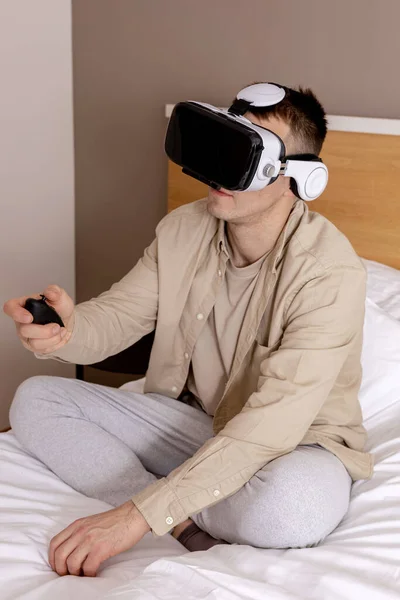VRヘッドセットを装着して自宅のベッドに座ってインタラクティブなビデオゲームをプレイし、仮想現実を探索する若い白人男性。VRメガネをかけた男。未来、ガジェット、技術、教育、オンライン学習. — ストック写真