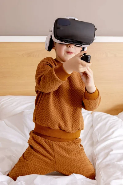 Маленький чарівний хлопчик сидить у ліжку вдома з гарнітурою VR і грає в інтерактивну відеогру, досліджуючи віртуальну реальність. Мила дитина в окулярах VR. Майбутнє, гаджети, технології, освіта онлайн . — стокове фото