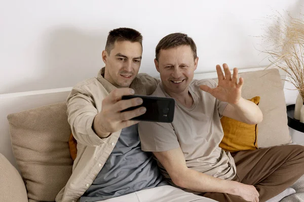 Šťastný gay pár s ležérní oblečení drží smartphone a mluvit s přáteli on-line, s videohovory. Homosexuální vztahy a alternativní láska. — Stock fotografie