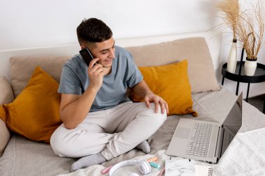 Gündelik kıyafetleri olan genç bir adam dizüstü bilgisayarı ve akıllı telefonuyla yatakta oturuyor. Freelancer evde çalışmak için defter kullanıyor. Mesafe çalışıyor, iş çevrimiçi.