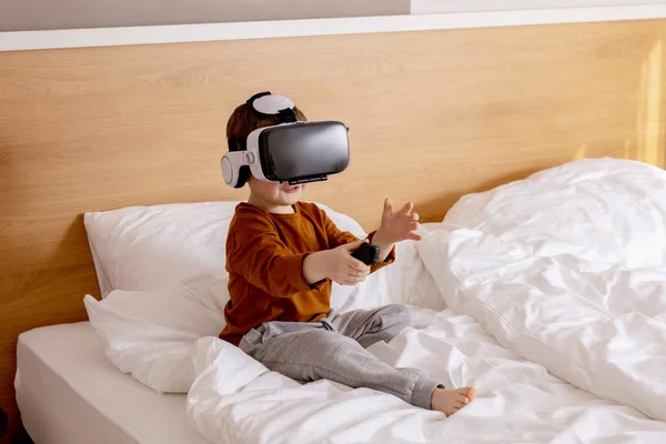 Little adorable boy sitting on bed at home with VR headset and playing interactive video game, exploring virtual reality. Cute child wearing VR glasses. Future, gadgets, technology, education online.