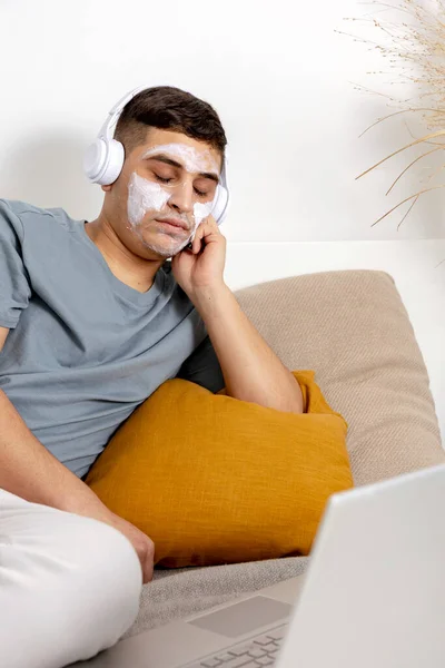 Mladý muž s neformálním oblečením a maskou krásy na obličeji ležící na posteli s notebookem a odpočívající. Kosmetika pro muže, péče o pleť. Muž poslouchající hudbu nebo audioknihu. Relaxace doma. — Stock fotografie