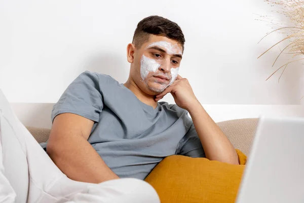 Mladý muž s neformálním oblečením a maskou krásy na obličeji ležící na posteli s notebookem a odpočívající. Kosmetika pro muže, péče o pleť. Člověk čte zprávy na internetu nebo se dívá na film. Relaxace doma. — Stock fotografie