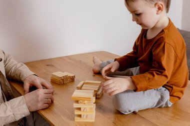 Baba ve oğul evde birlikte oturup tahta bloklarla oynuyorlar. Jenga oyunu. Küçük çocuk ve babası birlikte vakit geçiriyorlar. Aile zamanı. İçerde boş zaman aktivitesi.