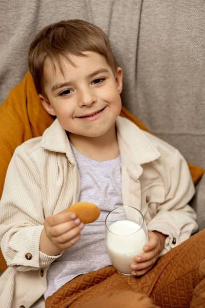 Little adorable boy sitting on the couch at home, drinking milk with cookie. Fresh milk in glass, dairy healthy drink. Healthcare, source of calcium, lactose. Preschool child with casual clothing. — Stock Photo, Image