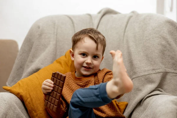 Niño adorable sentado en el sofá en casa y comiendo chocolate. Niño y dulces, confitería de azúcar. Los niños disfrutan de un delicioso postre. Niño en edad preescolar con ropa casual. Emoción positiva. — Foto de Stock