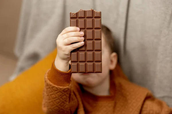 Piccolo adorabile ragazzo seduto sul divano a casa e mangiare barretta di cioccolato. Bambino e dolci, dolciumi. Il ragazzo si gode un delizioso dessert. Bambino in età prescolare con abbigliamento casual. — Foto Stock