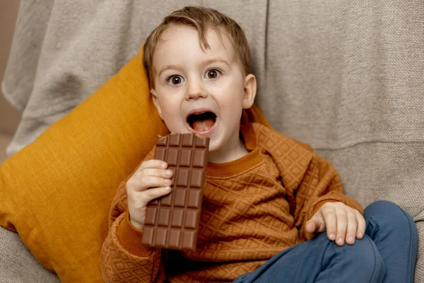 可爱的小男孩坐在家里的沙发上，吃着巧克力棒。孩子和糖果，糖糖果。孩子们享受美味的甜点.穿着休闲装的学龄前儿童。积极的情绪. — 图库照片