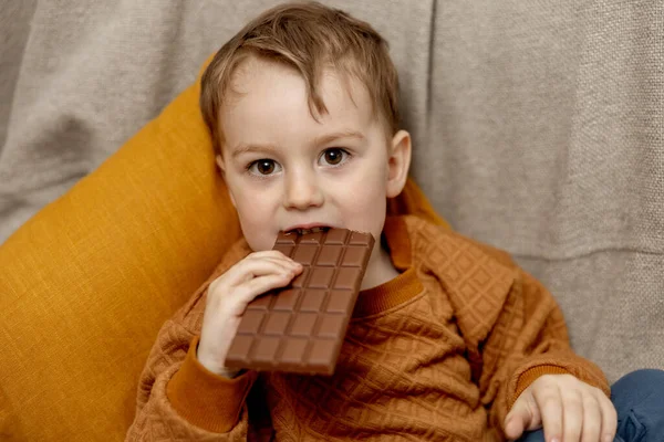 Petit garçon adorable assis sur le canapé à la maison et mangeant des barres de chocolat. Enfant et sucreries, sucreries. Les enfants apprécient un délicieux dessert. Enfant préscolaire avec des vêtements décontractés. Émotion positive. — Photo