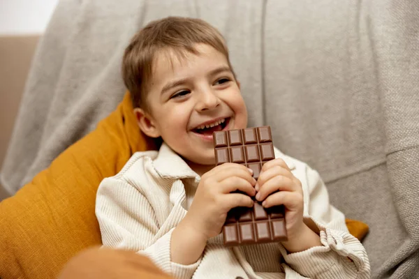 집에 있는 쇼파에 앉아 초콜릿 바를 먹는 귀여운 소년. 아이들 과 사탕, 설탕 과자. 애가 맛있는 디저트를 좋아하거든요. 평상복을 입은 취학 아동. 긍정적 인 감정. — 스톡 사진