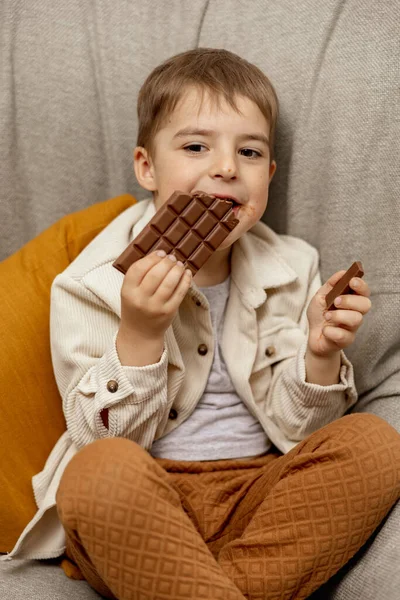 집에 있는 쇼파에 앉아 초콜릿 바를 먹는 귀여운 소년. 아이들 과 사탕, 설탕 과자. 애가 맛있는 디저트를 좋아하거든요. 평상복을 입은 취학 아동. 긍정적 인 감정. — 스톡 사진