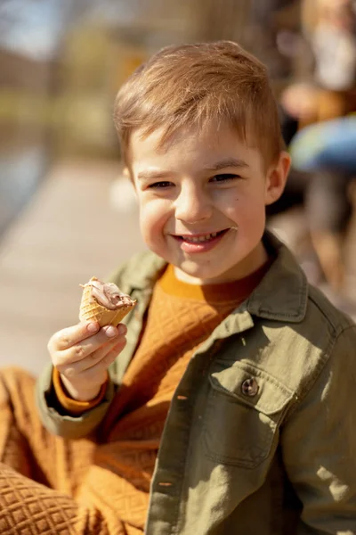 一个可爱的小男孩坐在户外吃着冰激凌。湖水和阳光普照的天气孩子和糖果，糖。孩子们享受美味的甜点.穿着休闲装的学龄前儿童。积极的情绪 — 图库照片