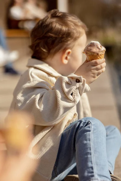 귀여운 소년 이 밖에 앉아서 아이스크림을 먹고 있었어요. 호수, 물, 그리고 화창 한 날씨. 아이와 사탕, 설탕. 애가 맛있는 디저트를 좋아하거든요. 평상복을 입은 취학 아동. 긍정적 인 감정 — 스톡 사진