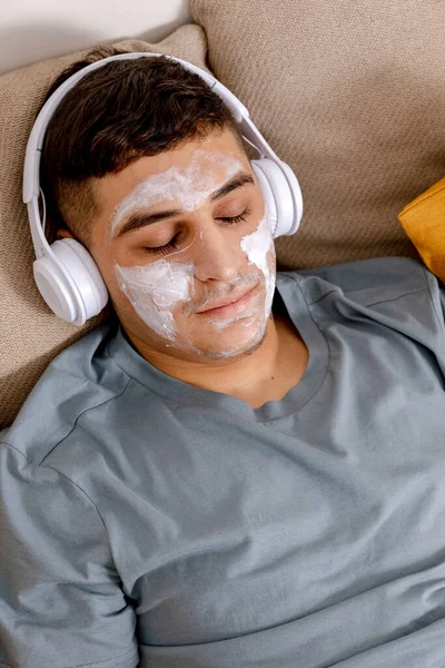 Young man with casual clothes and beauty mask on the face lying on bed at home and resting. Cosmetic for men, skin care. Man listening music or audio book. Relaxing. Time for yourself, mental health.