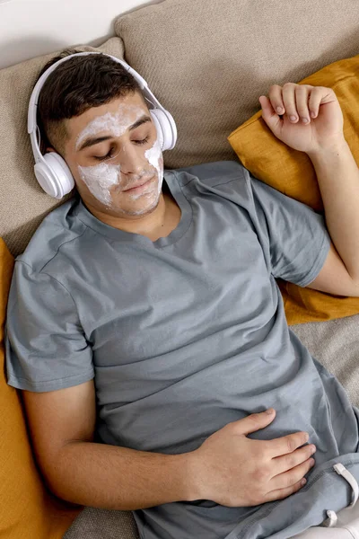 Young man with casual clothes and beauty mask on the face lying on bed at home and resting. Cosmetic for men, skin care. Man listening music or audio book. Relaxing. Time for yourself, mental health. — Stock fotografie