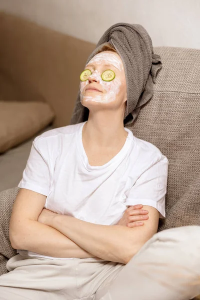 Ung kvinna med casual kläder, handduk på huvudet och skönhet mask i ansiktet sitter på soffan och vilar. Slappna av efter duschen. Kosmetisk för kvinnor, hudvård. Självhjälp, tid för dig själv. Avslappnande, — Stockfoto