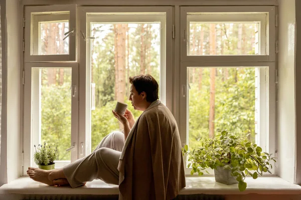 Middle age beautiful woman sitting on windowsill, drinking tea, dreaming. 50-year-old woman relaxing with cup of tea. Relax at home, looking away, thinking, stress free, peaceful mood wellbeing alone