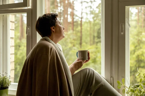 Middle age beautiful woman sitting on windowsill, drinking tea, dreaming. 50-year-old woman relaxing with cup of tea. Relax at home, looking away, thinking, stress free, peaceful mood wellbeing alone