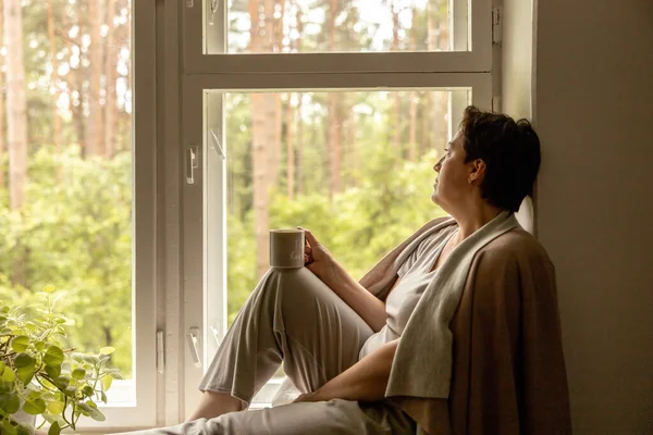 Middle age beautiful woman sitting on windowsill, drinking tea, dreaming. 50-year-old woman relaxing with cup of tea. Relax at home, looking away, thinking, stress free, peaceful mood wellbeing alone