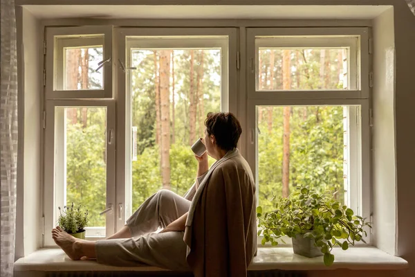 Middle age beautiful woman sitting on windowsill, drinking tea, dreaming. 50-year-old woman relaxing with cup of tea. Relax at home, looking away, thinking, stress free, peaceful mood wellbeing alone