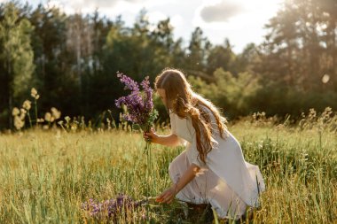 Genç, güzel bir kadın, beyaz bir elbise giyiyor, çayırda çiçekleri tutuyor. Doğanın ve özgürlüğün tadını çıkaran kız. Doğal güzellik. Stres yok, rüya görüyorsun. Gün batımı