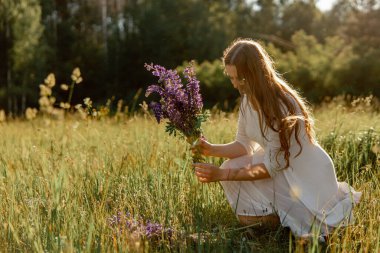 Genç, güzel bir kadın, beyaz bir elbise giyiyor, çayırda çiçekleri tutuyor. Doğanın ve özgürlüğün tadını çıkaran kız. Doğal güzellik. Stres yok, rüya görüyorsun. Gün batımı