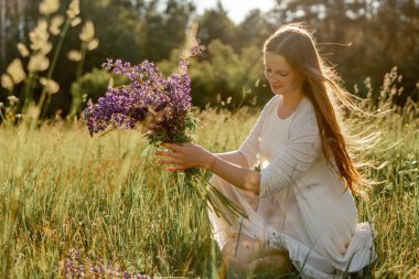 Genç, güzel bir kadın, beyaz bir elbise giyiyor, çayırda çiçekleri tutuyor. Doğanın ve özgürlüğün tadını çıkaran kız. Doğal güzellik. Stres yok, rüya görüyorsun. Gün batımı