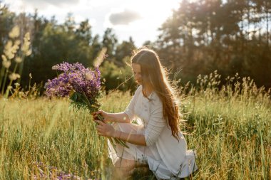 Genç, güzel bir kadın, beyaz bir elbise giyiyor, çayırda çiçekleri tutuyor. Doğanın ve özgürlüğün tadını çıkaran kız. Doğal güzellik. Stres yok, rüya görüyorsun. Gün batımı