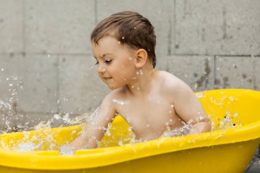 Cute little boy bathing in yellow tub outdoors. Happy child is splashing, playing with water and having fun. Summer season and recreation. Staying cool in the summer heat. Water fun in backyard