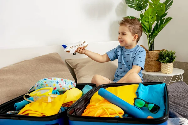 Little caucasian boy with blue shirt ready for vacation. Happy child packs clothes into a suitcase for travel. Tourist, joy of holiday. Kid at home, preparing for flying. Modern and cozy interior