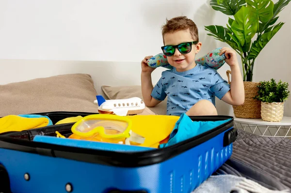 Little caucasian boy with blue shirt ready for vacation. Happy child packs clothes into a suitcase for travel. Tourist, joy of holiday. Kid at home, preparing for flying. Modern and cozy interior