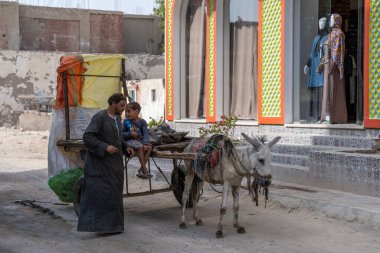 Hurghada, Mısır, 3 Kasım 2021: Eşeğe koşum takılmış bir araba yerel bir giyim mağazasının yanındaki engebeli bir yolda duruyor. Kirli, pis bir Arap çocuk arabada oturuyor, bir adam dizginleri almak için eşeğe gidiyor. Hayvan zulmü. Çöp toplayıcıları.