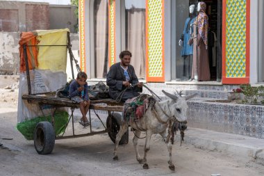 Hurghada, Mısır, 3 Kasım 2021: Eşek, kirli, kirli bir Arap ve çocuğun oturduğu bir araba taşıyor, çöp toplayıcılar bozuk bir yol boyunca yerel bir giyim mağazasının önünden geçiyor. Hayvan zulmü..