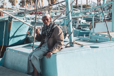 Hurghada, Mısır, 19 Kasım 2021: Rıhtımdaki balıkçı teknelerinin arka planına karşı geleneksel Arap elbisesi giymiş yaşlı bir balıkçının portresi. Yaşlı adam dişsiz bir ağızla teknelerden birinin kenarına yaslanarak gülümsüyor.