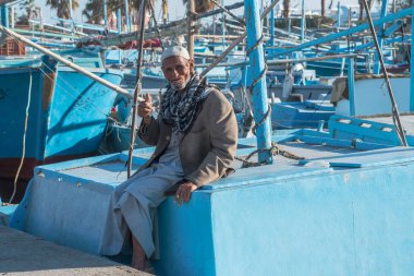 Hurghada, Mısır, 19 Kasım 2021: Rıhtımdaki balıkçı teknelerinin arka planına karşı geleneksel Arap elbisesi giymiş yaşlı bir balıkçının portresi. Yaşlı adam dişsiz bir ağızla teknelerden birinin kenarına yaslanarak gülümsüyor.