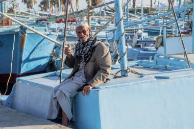 Hurghada, Mısır, 19 Kasım 2021: Rıhtımdaki balıkçı teknelerinin arka planına karşı geleneksel Arap elbisesi giymiş yaşlı bir balıkçının portresi. Yaşlı adam dişsiz bir ağızla teknelerden birinin kenarına yaslanarak gülümsüyor.