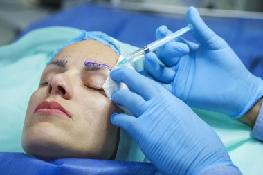 Kaş düzeltmesi için estetik ameliyat. Kadın hasta ameliyat masasında yatıyor. Doktor ameliyattan önce anestezi iğnesi yapıyor.