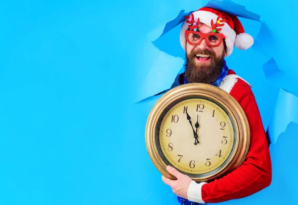 Новогодний отсчет. Улыбающийся Санта-Клаус со старинными часами смотрит сквозь бумажное отверстие. Готовлюсь к Рождеству. Копирование пространства, — стоковое фото