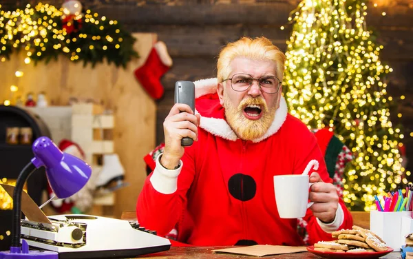 Θυμωμένος Άγιος Βασίλης κάθεται στην πολυθρόνα βλέποντας Χριστουγεννιάτικη ταινία και πίνει καφέ. Πρωτοχρονιάτικο σινεμά. — Φωτογραφία Αρχείου