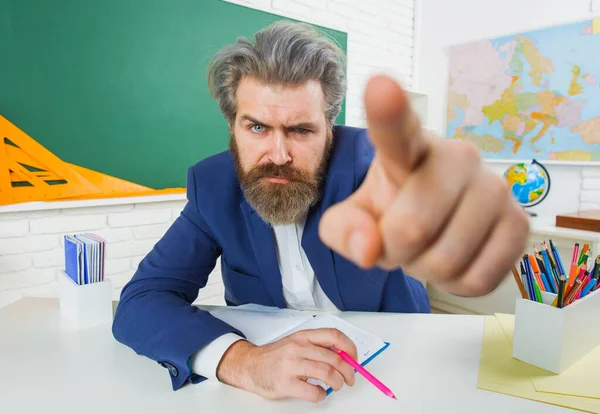 Θυμωμένος δάσκαλος στην τάξη που δείχνει με το δάχτυλο. Σοβαρός γενειοφόρος άνδρας με κοστούμι κάθεται στο τραπέζι στην τάξη. — Φωτογραφία Αρχείου