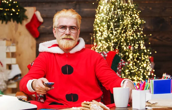 Санта Клаус сидит в кресле и смотрит рождественский фильм с пультом дистанционного управления. Новогодний кинотеатр дома. — стоковое фото