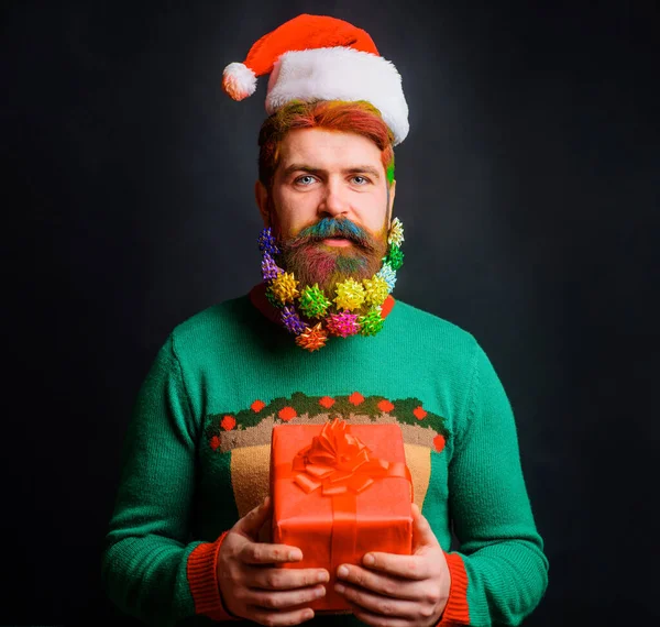 Бородатый мужчина в шляпе Санты с рождественским подарком. Хипстер с украшенной бородой к новогоднему празднику. — стоковое фото
