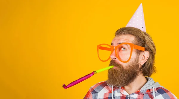 Homem barbudo de chapéu de festa faz barulho. Cara feliz em óculos grandes em boné de cone de aniversário. Espaço de cópia. — Fotografia de Stock