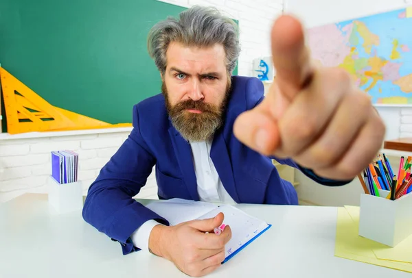 Σοβαρός δάσκαλος στην τάξη που δείχνει με δάχτυλο. Γενειοφόρος άνδρας κάθεται στο τραπέζι στην τάξη. Μάθηση, εκπαίδευση. — Φωτογραφία Αρχείου