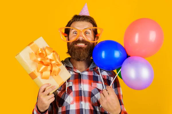 Feliz chico guapo celebrando cumpleaños. Hombre barbudo en gorra de vacaciones con globos y caja de regalo. — Foto de Stock
