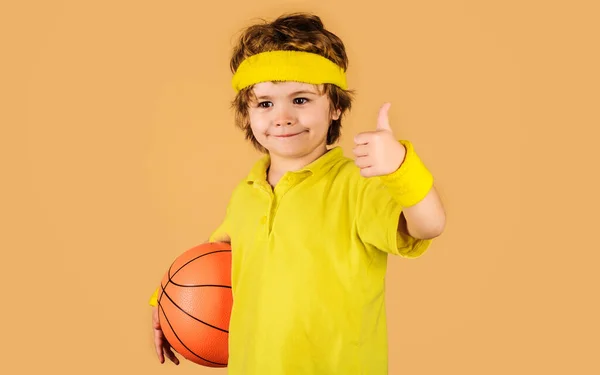 Niño deportivo con pelota de baloncesto mostrando el pulgar hacia arriba. Pequeño jugador de baloncesto en ropa deportiva. —  Fotos de Stock