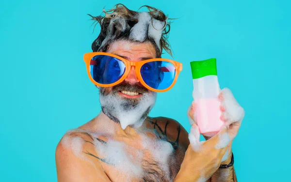 Grappige man onder de douche met shampoo fles. Een man met een baard en een grote bril met schuim op zijn hoofd. Haarspoeling. — Stockfoto